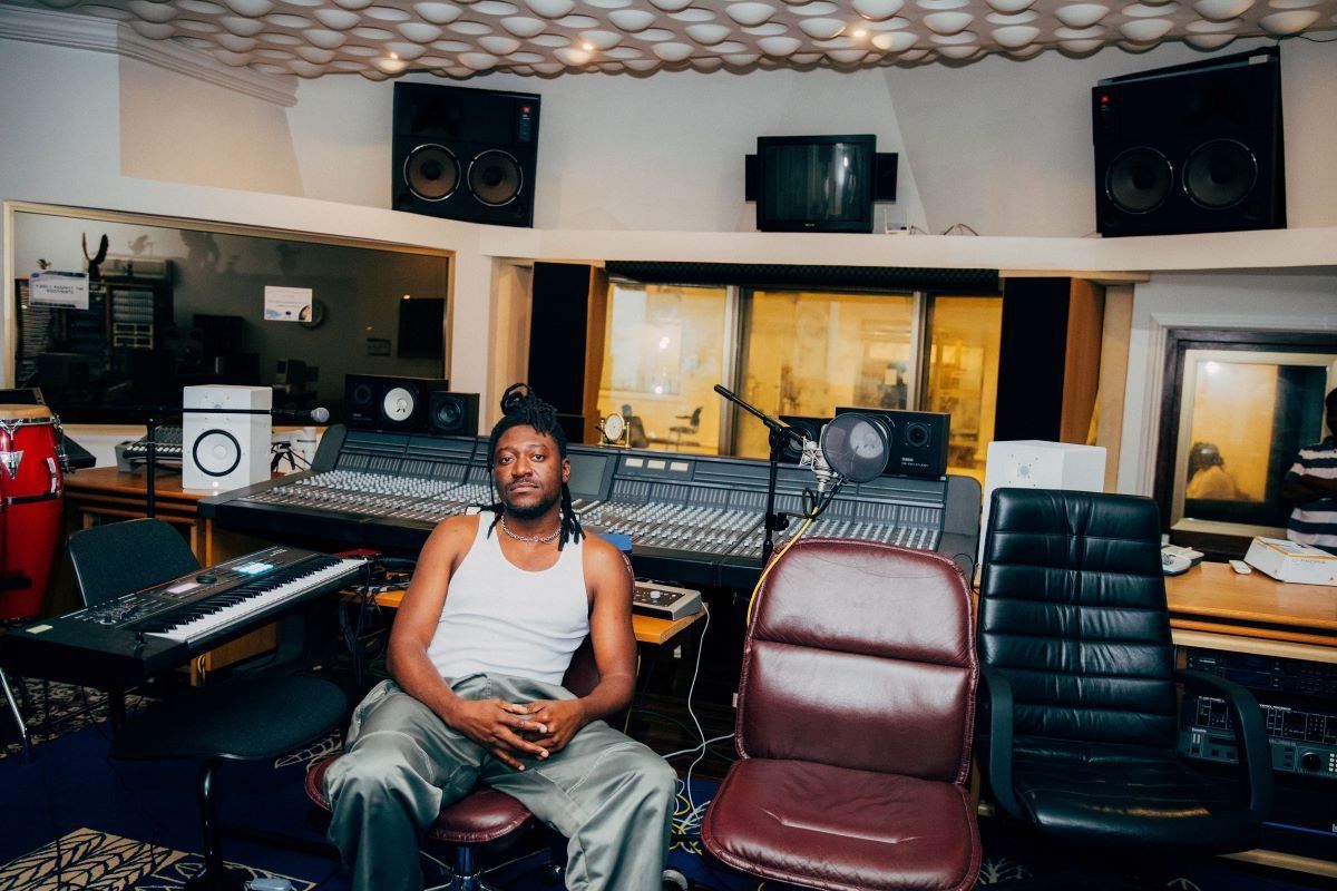 A photo of GuiltyBeatz sitting in a studio wearing white singlet and silver baggy pants.