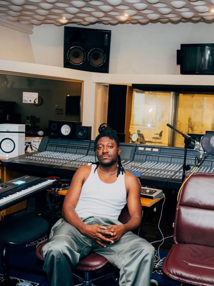A photo of GuiltyBeatz sitting in a studio wearing white singlet and silver baggy pants.