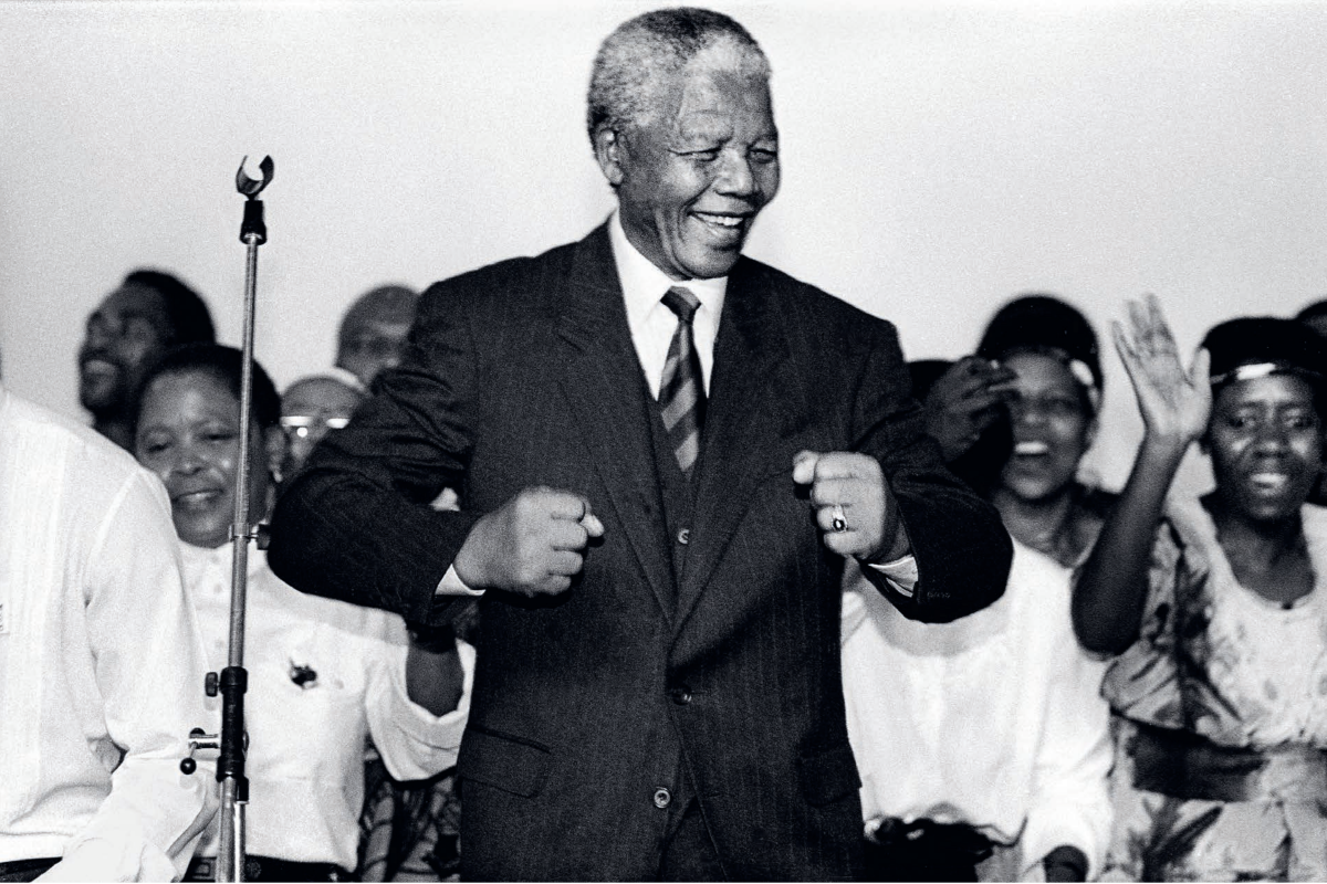 A photo of Nelson Mandela wearing a suit while smiling as he does his famous “Madiba shuffle” which involves forming a double fist and raising both arms in a curved position while moving the body slightly.