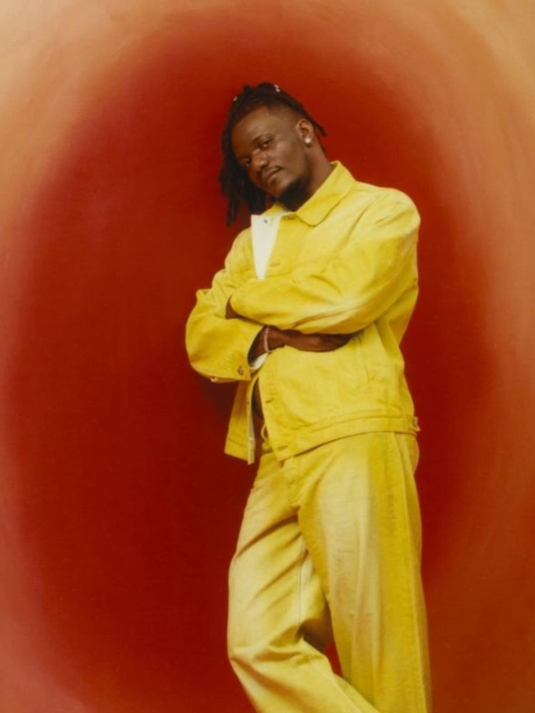 A photo of Pheelz resting his shoulders on an orange-coloured wall with arms crossed.