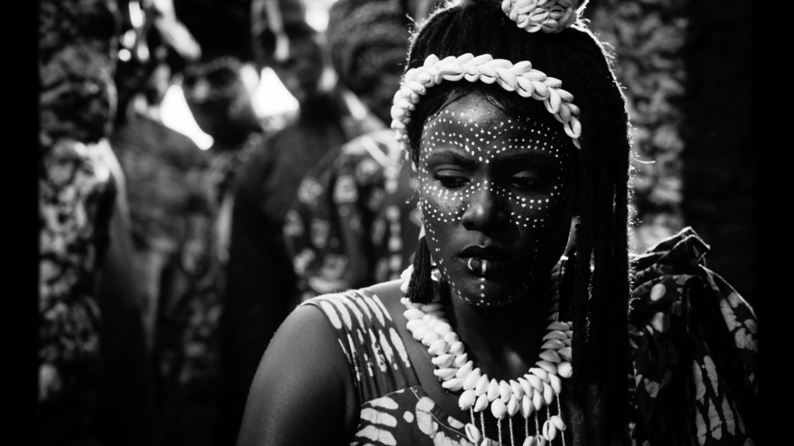 A black and white still from the film, Mami Wata, directed by CJ Obasi of a woman with Yoruba face paint on, looking with her eyes downcast.