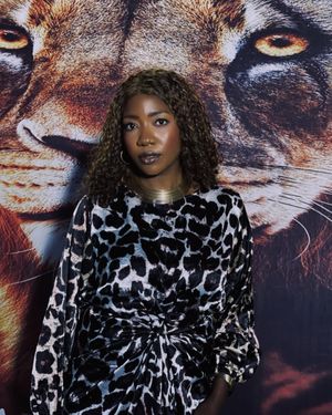 A Black woman with medium brown curly hair poses in front of a backdrop featuring a close-up of a lion's face.