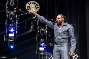 A man stands on a stage, wearing a denim jacket and pants while holding up a hand fan.