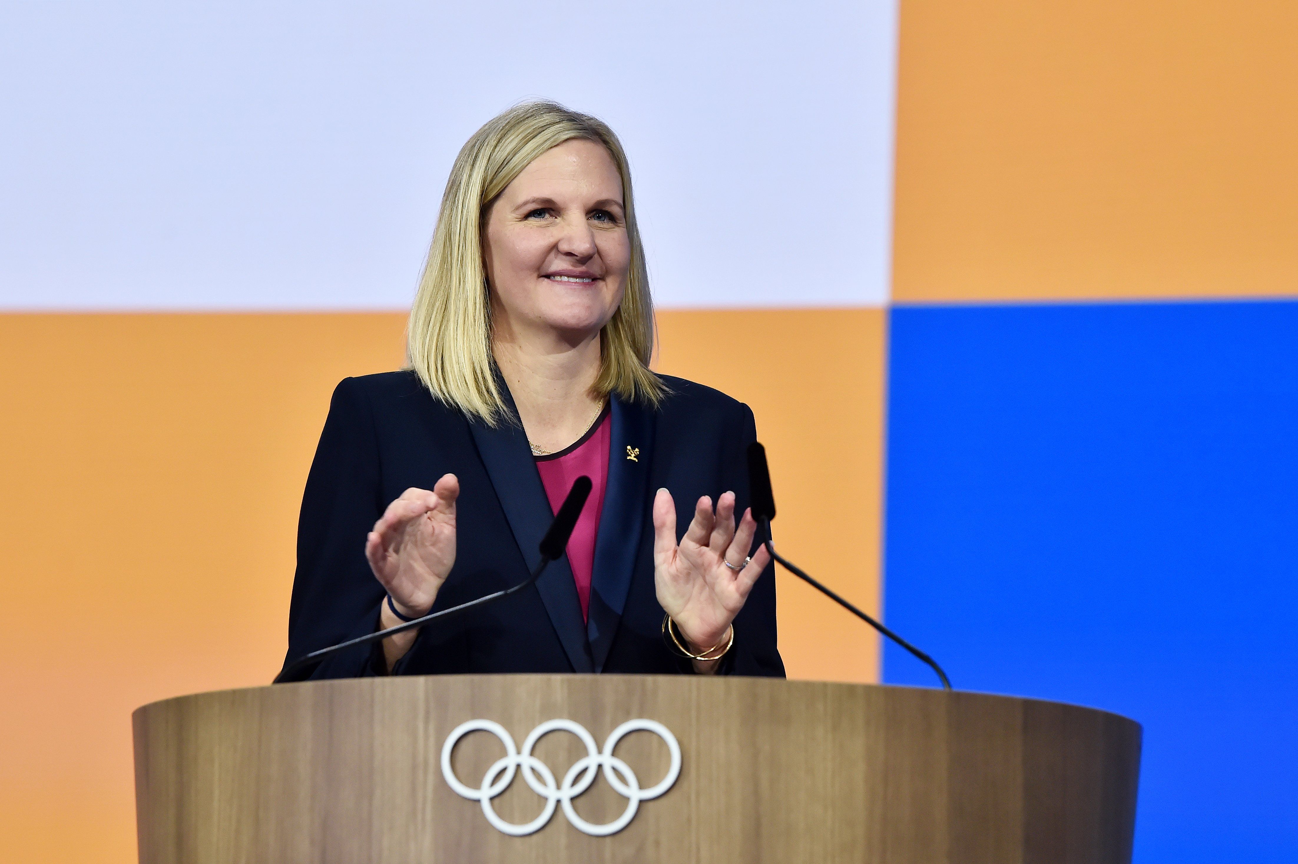 Kirsty Coventry smiles as she stands behind a wooden podium with the Olympic rings emblem on the front. There are two microphones on the podium and she is delivering a speech, gesturing with her hands, which are positioned in front of her, slightly apart, with open palms. She has shoulder-length blonde hair, and is wearing a navy-blue blazer over a burgundy top.