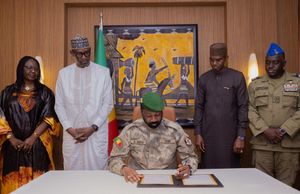 Military official wearing green beret and camouflage uniform signs documents at desk while four officials stand behind, with Malian flag and African artwork in background.