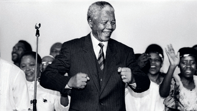 A photo of Nelson Mandela wearing a suit while smiling as he does his famous “Madiba shuffle” which involves forming a double fist and raising both arms in a curved position while moving the body slightly.
