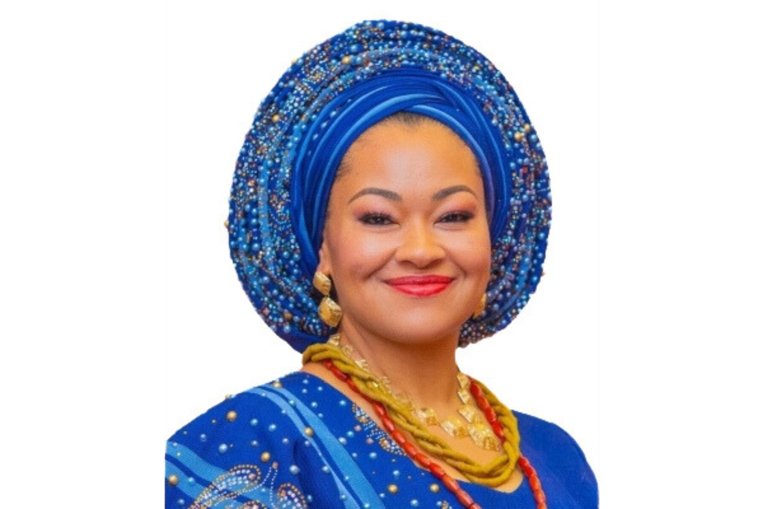 Senator Natasha Akpoti in a beaded blue traditional Nigerian outfit with a matching blue head wrap (gele), gold earrings, colorful necklaces, and red lipstick, smiling at the camera.