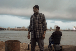 'The Black Book' actor Richard Mofe-Damijo standing on a shoreline under a cloudy sky with Alex Usifo in a wheelchair facing the water.