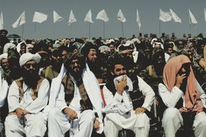 men in middle eastern dress seated in chairs with white flags containing Arabic text flying in the background.